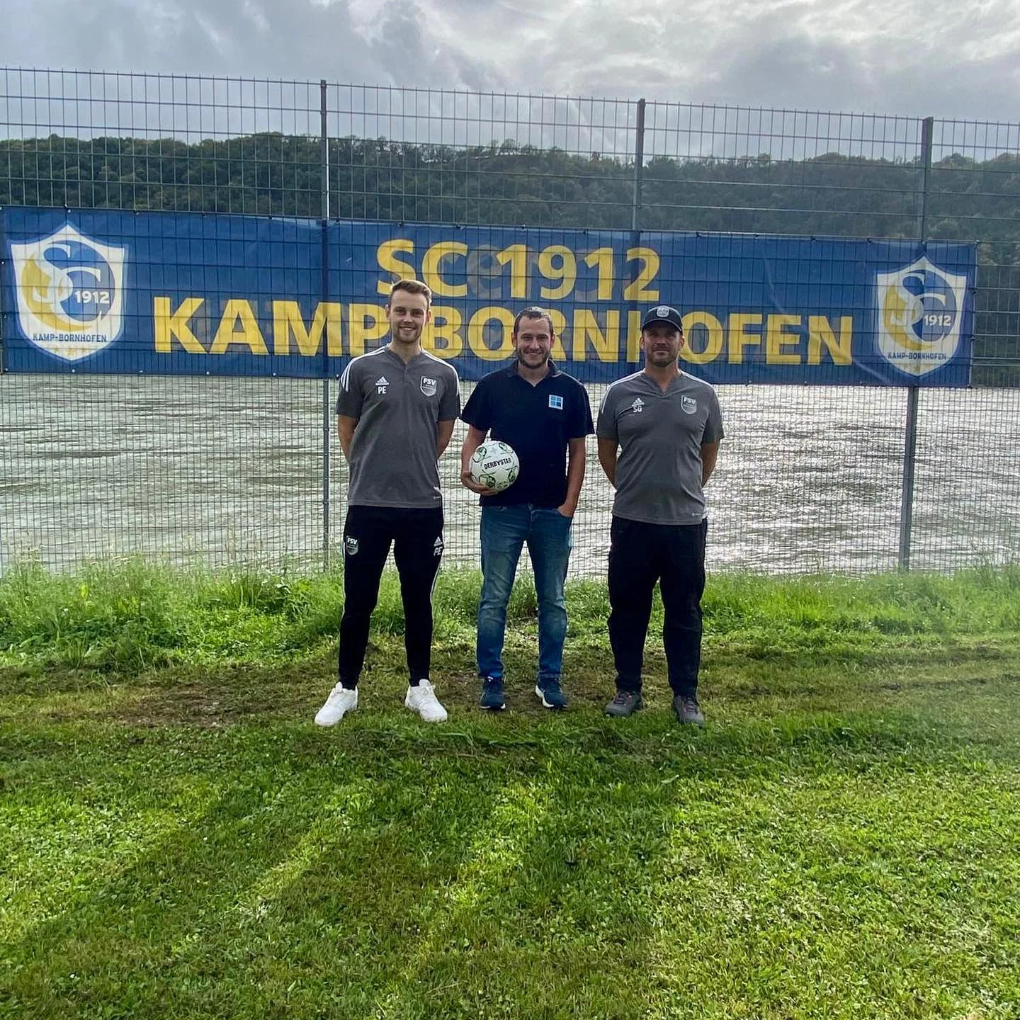 Übergabe der neuen Fußbälle vor dem Banner des SC 1912 Kamp-Bornhofen: drei Personen auf dem Sportplatz, in der Mitte ein Vertreter von Technik-Schneider mit einem der neuen Bälle